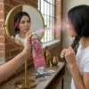Una mujer joven frente a un espejo circular aplicando el spray de color rosa en una sección de su cabello trenzado. Se ve el reflejo de su rostro mientras rocía el producto en un ambiente luminoso con paredes de ladrillo.