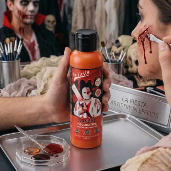 Un maquillador profesional de efectos especiales creando una herida realista en el rostro de una mujer con una espátula y sangre decorativa viscosa. Sostiene la botella del producto. Un plato de metal con muestras de sangre y un cartel que dice "LA FIESTA: ARTISTRY IN EFFECTS" están sobre la mesa.