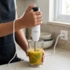 Persona usando la batidora de mano Kiwi para triturar verduras y frutas en un vaso medidor, logrando una textura suave y sin grumos.