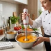 Chef utilizando batidora de mano para triturar una crema de verduras naranja en un bol de acero inoxidable.