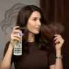 Mujer de cabello castaño aplicando spray de color marrón para retocar raíces y dar brillo instantáneo al peinado.