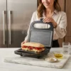 Mujer preparando un sándwich de jamón y pimientos en una prensa de panini eléctrica en una cocina moderna.