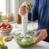 Persona utilizando la batidora de mano Kiwi para batir una salsa de albahaca y piñones en un bol de cristal, demostrando su facilidad de uso.