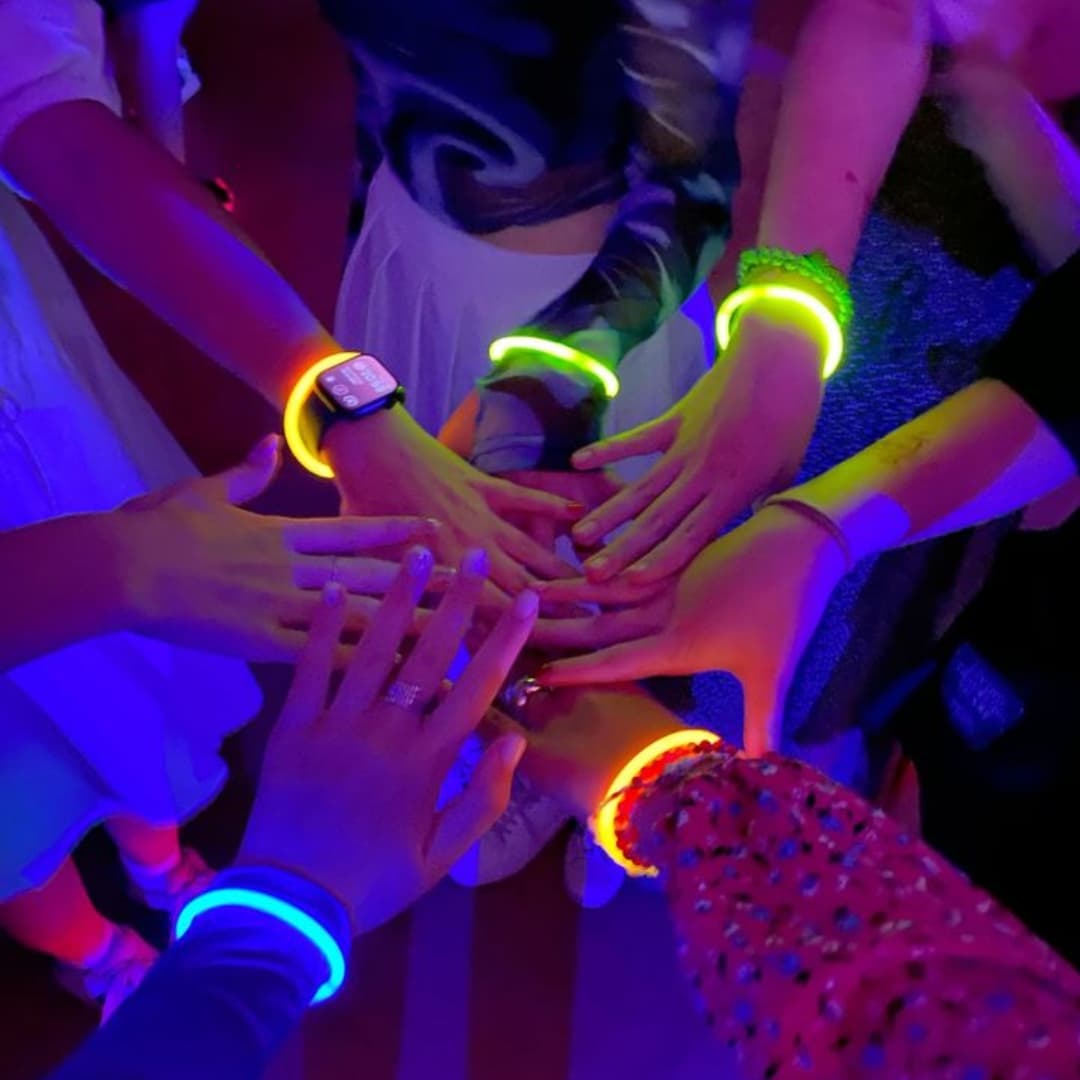 Manos juntas con pulseras luminosas Glow Elite encendidas mostrando colores intensos en la oscuridad