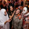 Maquillaje Zombie Realista en Fiesta de Halloween Una mujer vestida de vampiresa aplicando sangre decorativa viscosa en la mejilla de un hombre disfrazado de zombie, quien sonríe y sostiene la botella del producto. Un fantasma y un personaje con máscara de hockey observan.