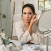 Mujer con albornoz beige aplicando una pestaña postiza azul eléctrico con pinzas doradas frente a un espejo de aumento en un baño iluminado.